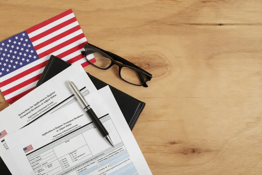 A flat lay image of immigration application forms, a pen, glasses, and a small U.S. flag.